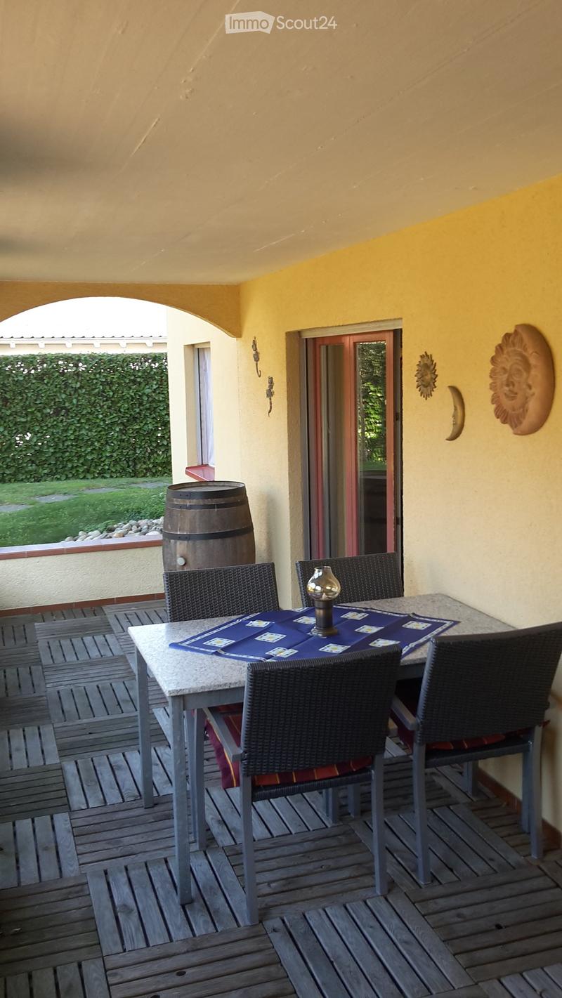 Wooden deck with a round table, four wicker chairs, and a small lamp in the center. The table is covered with a blue placemat and has playing cards on it. There is a large barrel in the background and a view of a garden through an open doorway.