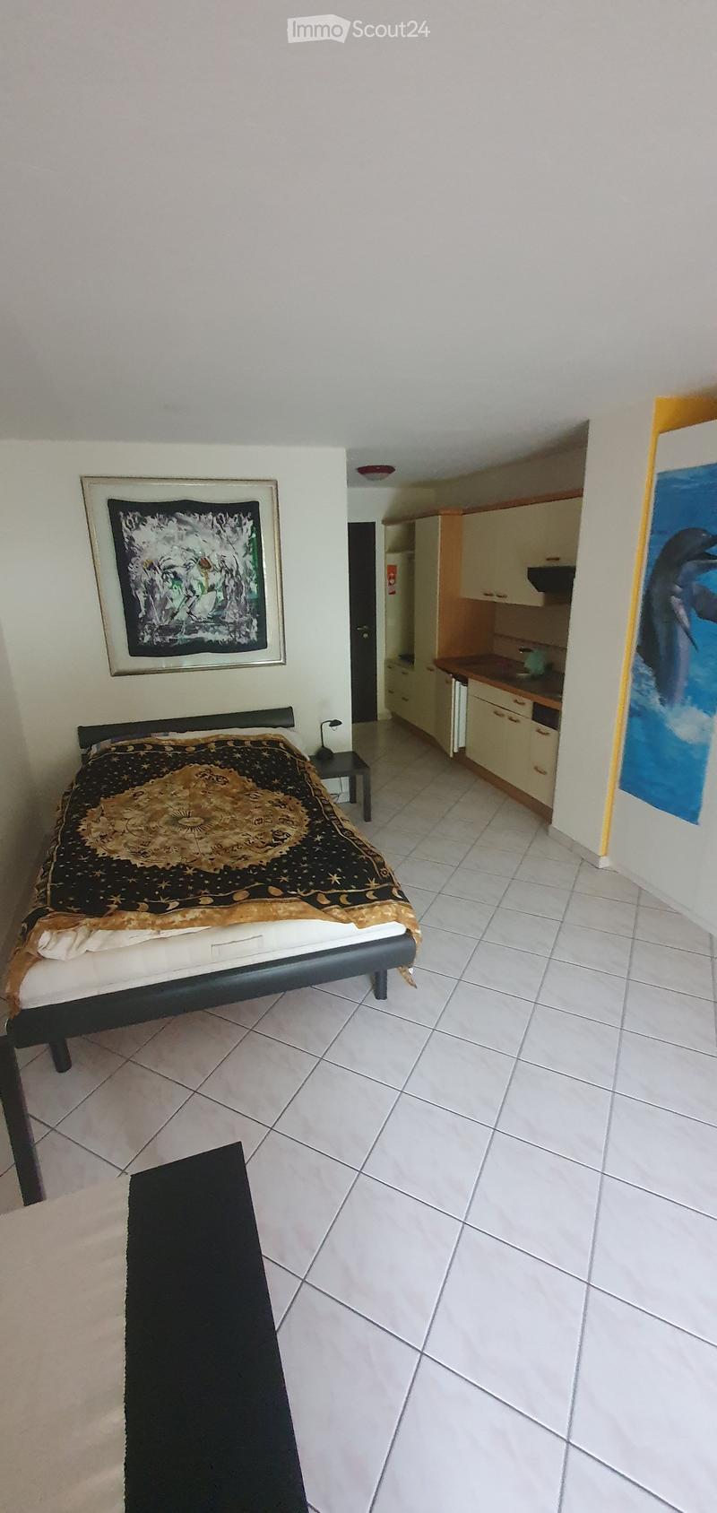 A furnished bedroom with a bed, nightstand, and a small table, connected to a compact kitchen area with cabinets and a counter.