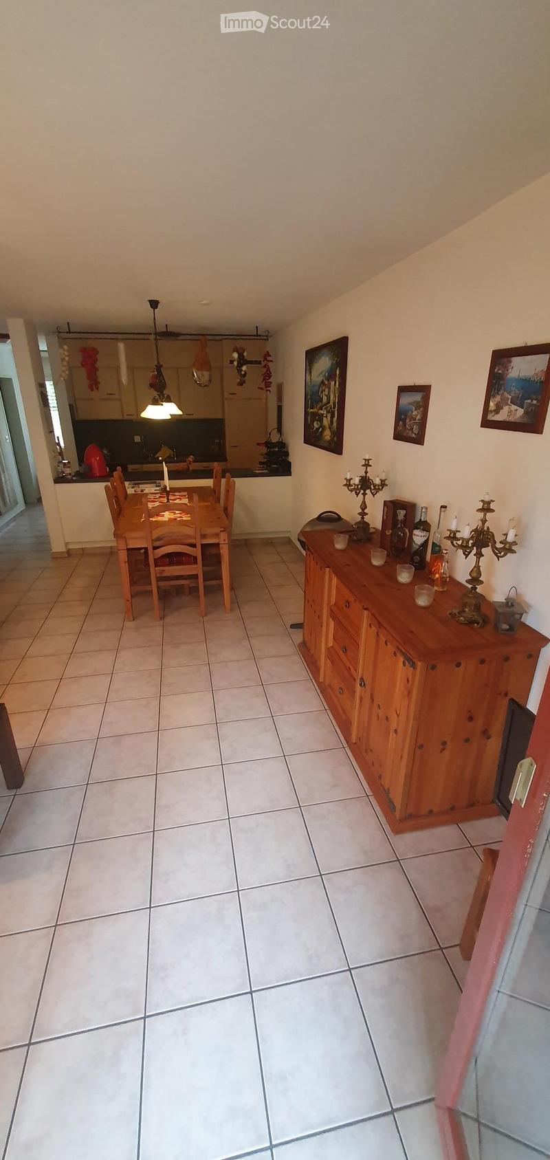 Dining area with a wooden dining table, chairs, a wooden cabinet with drawers, and decorative items including candle holders and picture frames. The room has tiled flooring and is connected to a kitchen area visible in the background.