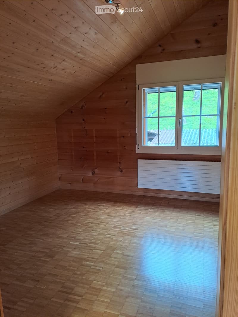 Empty attic room with wooden walls, wooden floor, large window, and radiator.
