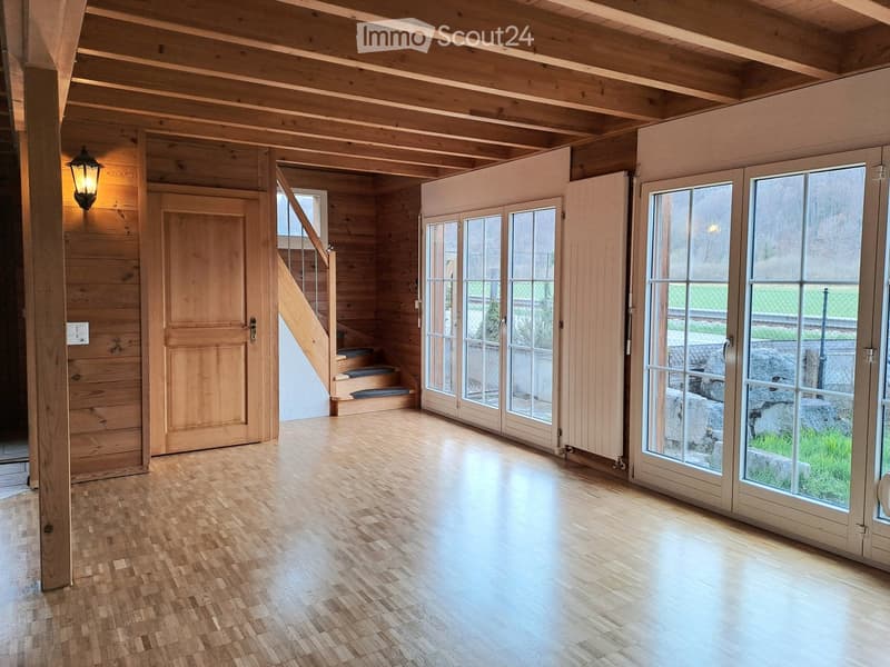Bright, empty room with wooden floors and exposed wooden beams. Features large glass doors leading outside, a staircase to an upper level, and a wall-mounted light fixture.