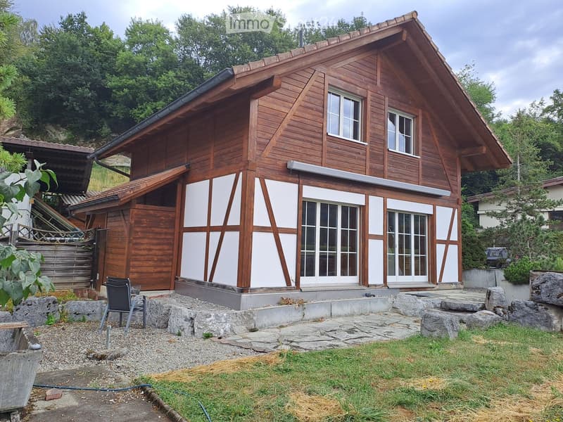 Two-story wooden house with white trim, large windows, and a garage. The house is surrounded by greenery and stone pathways.
