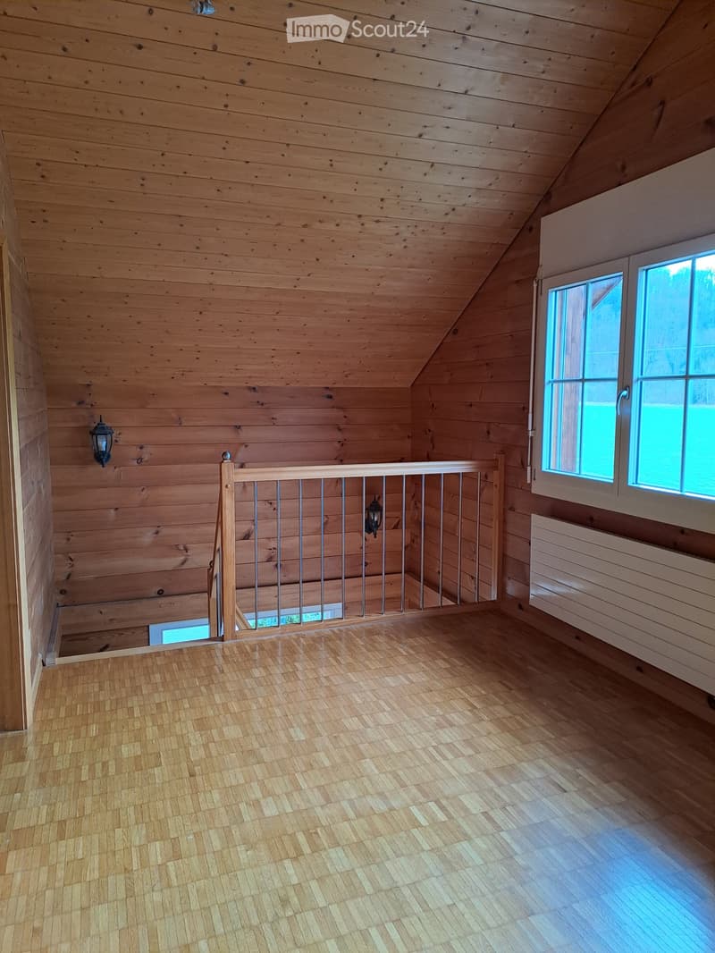 Empty attic space with wooden walls, ceiling, and floor, featuring a metal railing and two windows letting in natural light.