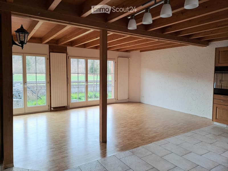 Empty room with wooden floor, exposed wooden ceiling beams, large windows, and a small kitchen area with cabinets.
