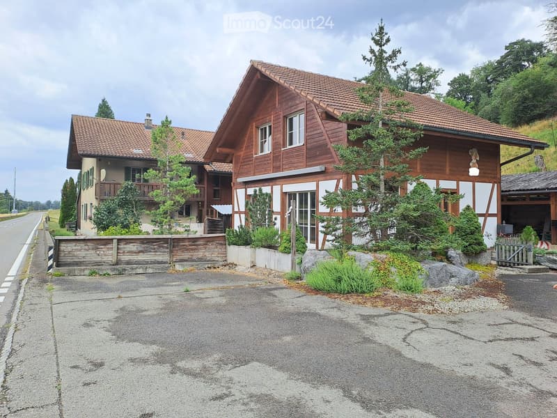 Two-story wooden house with red-tiled roof, multiple windows, and a small balcony on the left side. The house is surrounded by greenery and a wooden fence.