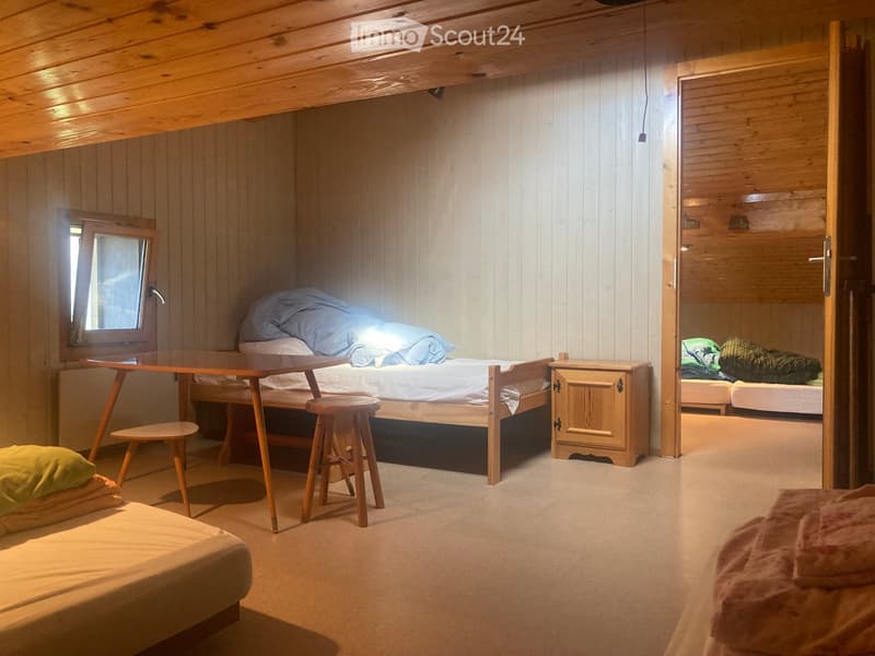 Cozy wooden bedroom with multiple beds, a small table, a stool, and a mirror. The room features wooden walls and a slanted ceiling.