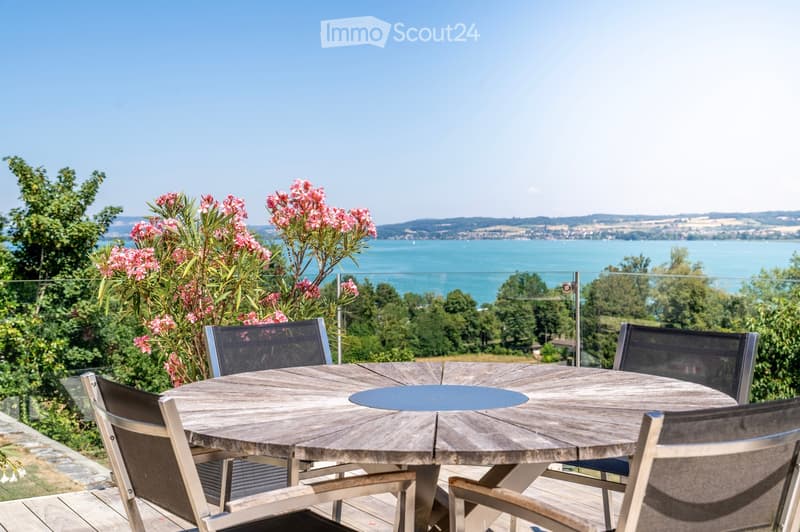 Balcony with a round wooden table and four chairs, overlooking a scenic lake and distant hills.