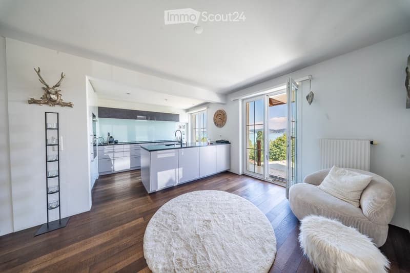 Open-plan living and dining area with a modern kitchen, white cabinets, a central island, a round white rug, a beige sofa, and a decorative wall piece.