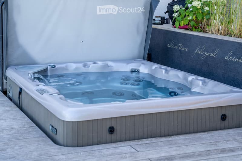 A modern hot tub with multiple jets, placed on a wooden deck, surrounded by a grey wall with decorative text and a small potted plant.