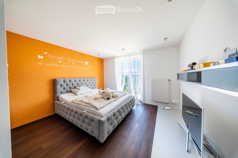 Bright bedroom with wooden flooring, a large window with white curtains, a grey upholstered bed with pillows and a teddy bear, a radiator, and a cabinet with a remote control on the floor.