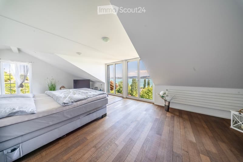 Bright bedroom with wooden flooring, large windows offering a scenic view, a neatly made bed, a nightstand, and decorative vases.