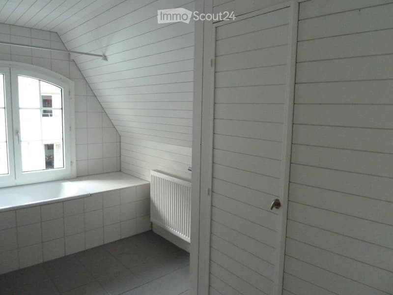 An unfurnished attic room with white wooden panel walls, a slanted ceiling, a window, and a radiator. The room is empty with no furniture or personal items.