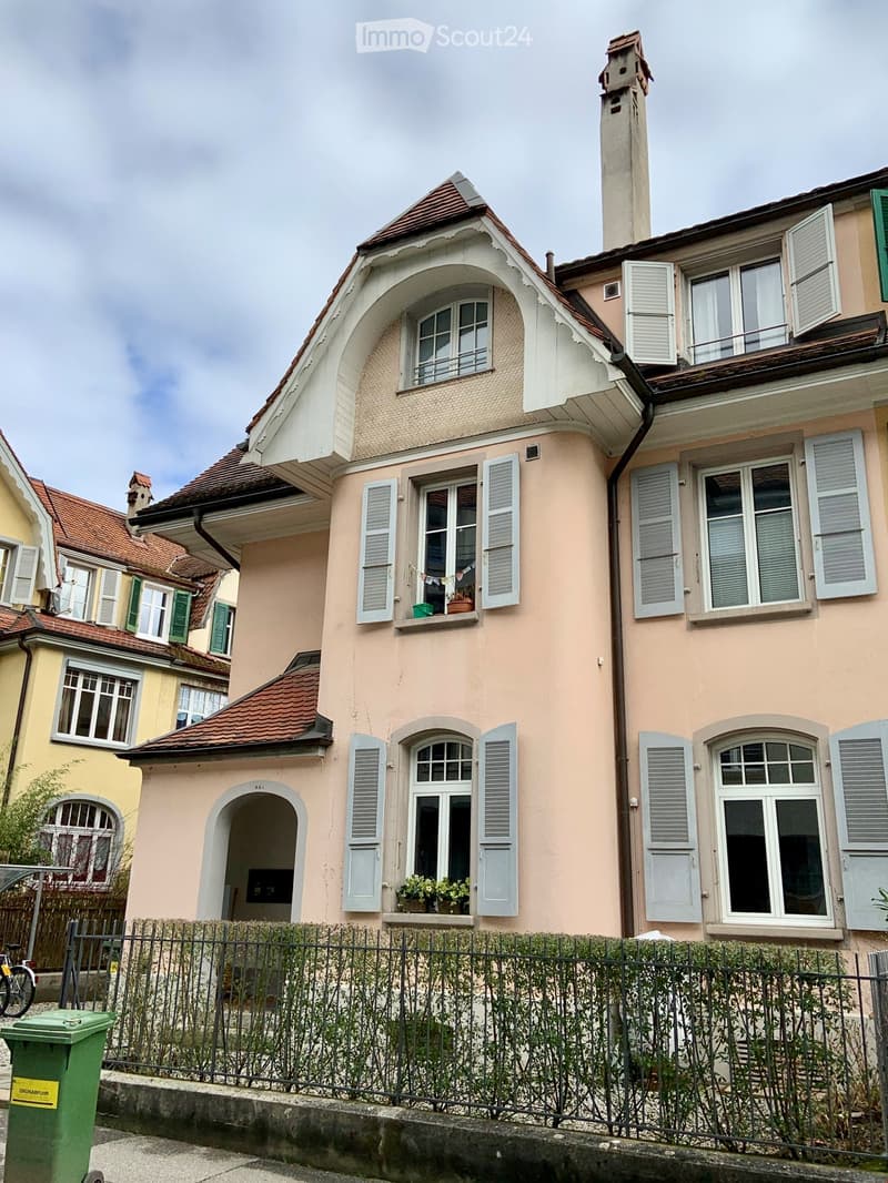 Two-story house with pink facade, multiple windows with shutters, decorative roofline, and small balcony with potted plants.