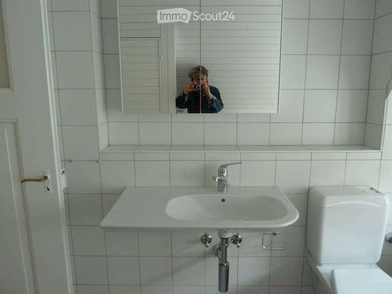 A modern bathroom with white tiled walls and floor, featuring a white sink with a chrome faucet, a toilet, and a large mirror with a built-in shelf above the sink.