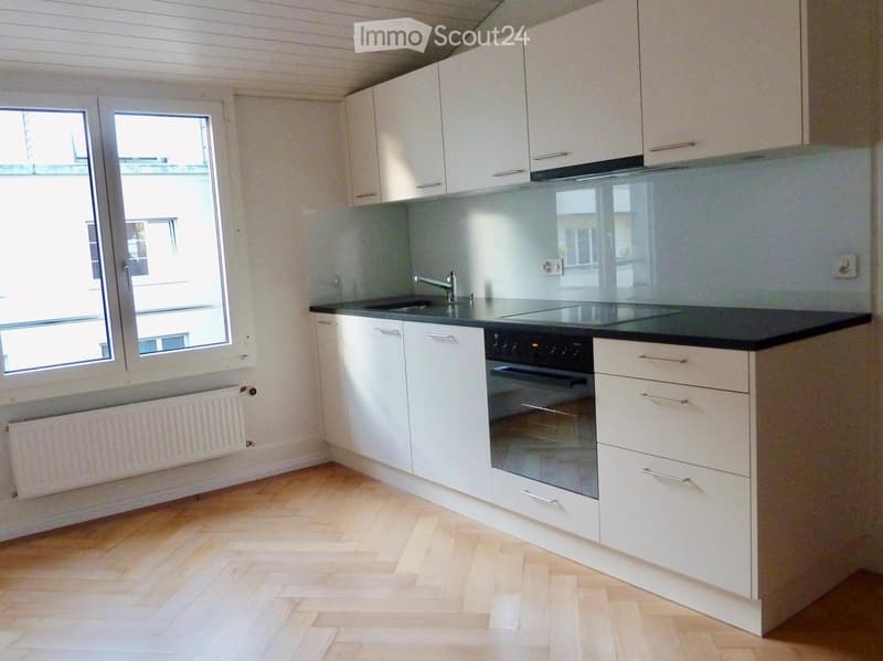 Modern kitchen with white cabinets, black countertop, sink, faucet, and built-in oven. Light wooden flooring and large windows provide natural light.