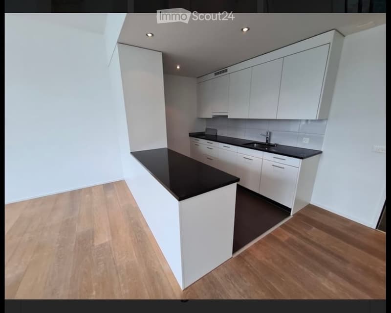 Modern kitchen with white cabinets, black countertops, and a central island. The floor is wooden, and the walls are white.