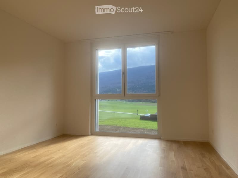 Empty room with wooden flooring and large windows offering views of a green field and mountains.
