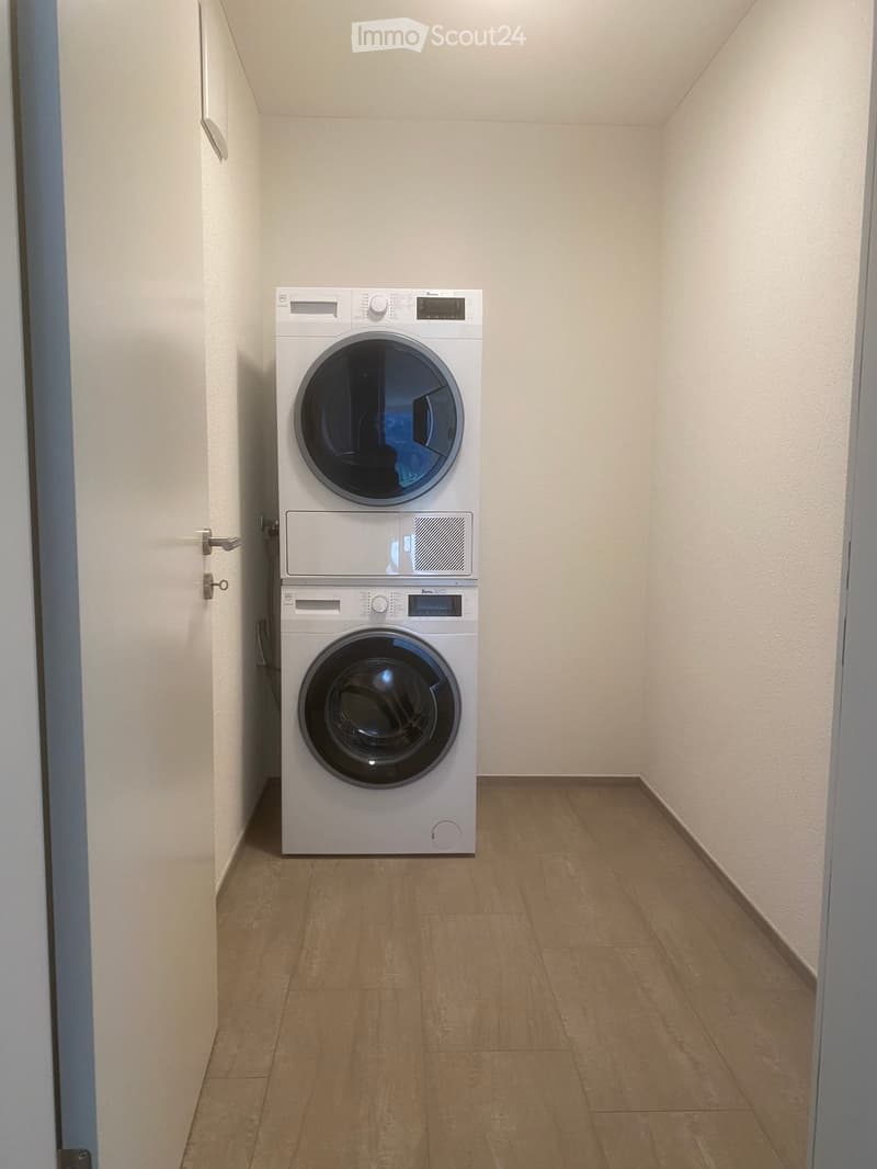 A small, well-lit room with light-colored walls and wooden flooring, featuring a stacked washing machine and dryer unit.
