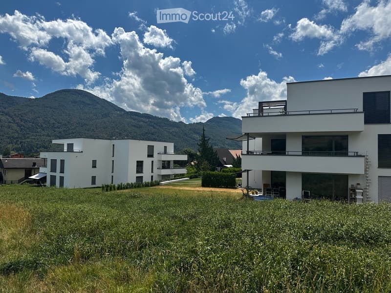 Modern two-story white house with large windows and balconies, surrounded by a grassy field and mountains in the background.