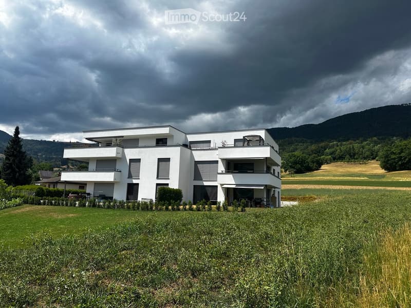 Modern two-story white house with large windows, balconies, and a spacious lawn in front, surrounded by greenery and hills in the background.