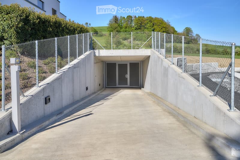 Modern underground garage entrance with concrete walls, metal fence, and a raised ground floor.