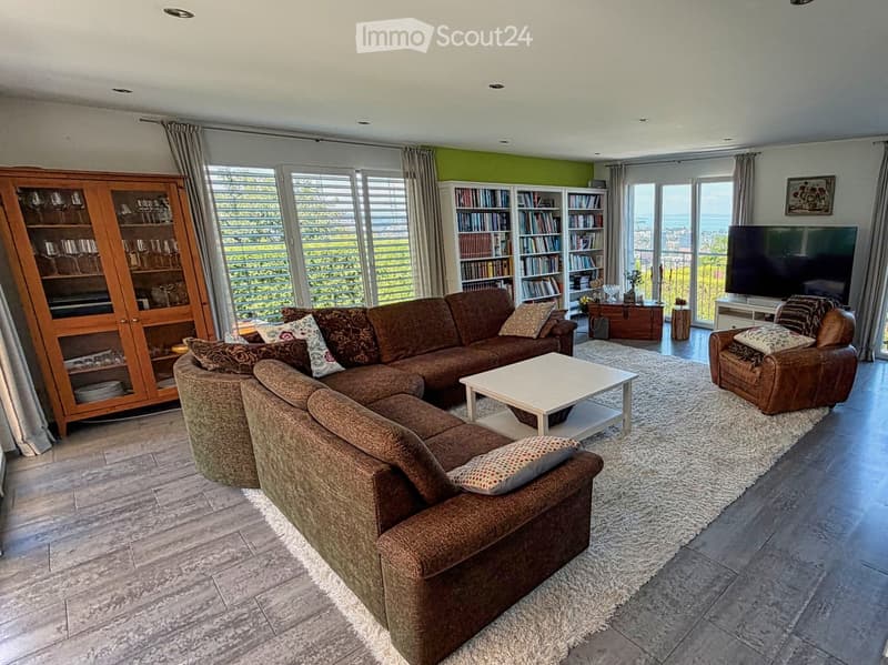 Modern living room with large windows offering a scenic view, featuring a brown sectional sofa, a white coffee table, an armchair, a bookshelf, a cabinet, and a television.