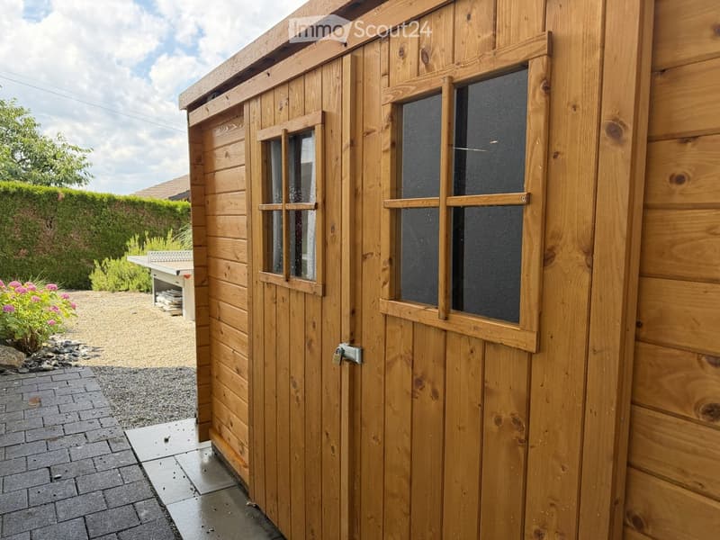 A wooden shed with two windows and a door, situated on a paved area with gravel and a small garden nearby.