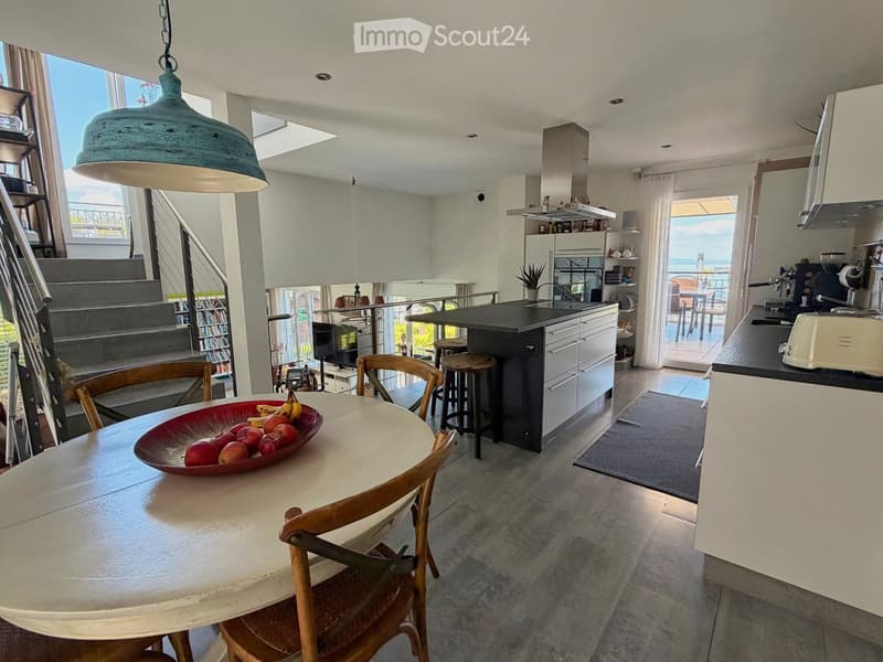 Open-plan kitchen and dining area with a round wooden table, chairs, stools, modern appliances, and a view of the staircase and balcony.