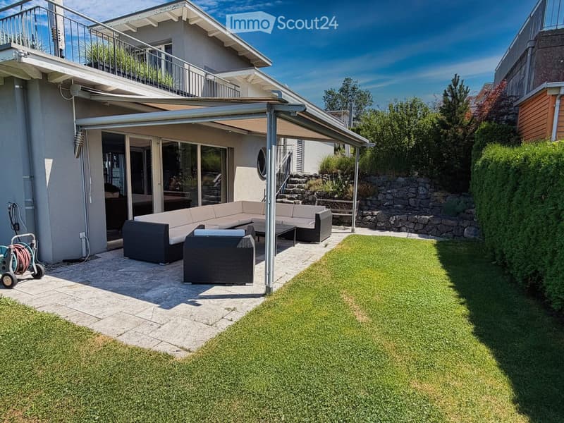 Outdoor patio with a modern sofa set, a wicker chair, and a small table, covered by a retractable awning, adjacent to a well-maintained lawn and a stone wall.