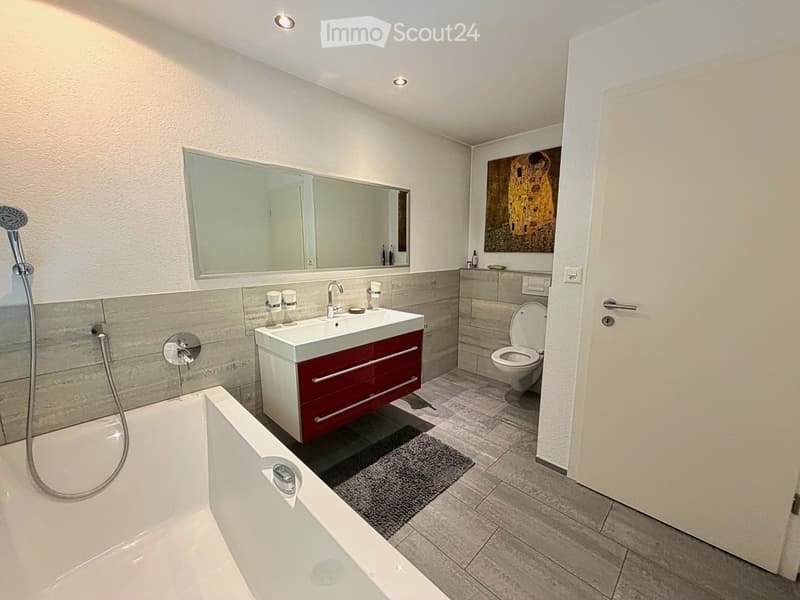 Modern bathroom with a bathtub, sink, toilet, large mirror, and a showerhead. The sink has a red cabinet underneath, and the floor is tiled.