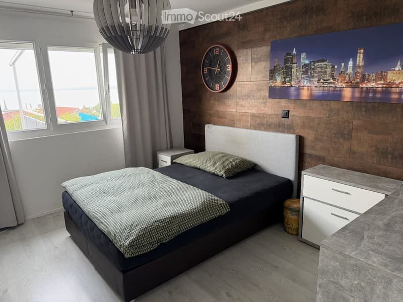 Modern bedroom with a double bed, nightstand, wall-mounted clock, and large cityscape picture on the wall. The room features wooden flooring and large windows with a view.