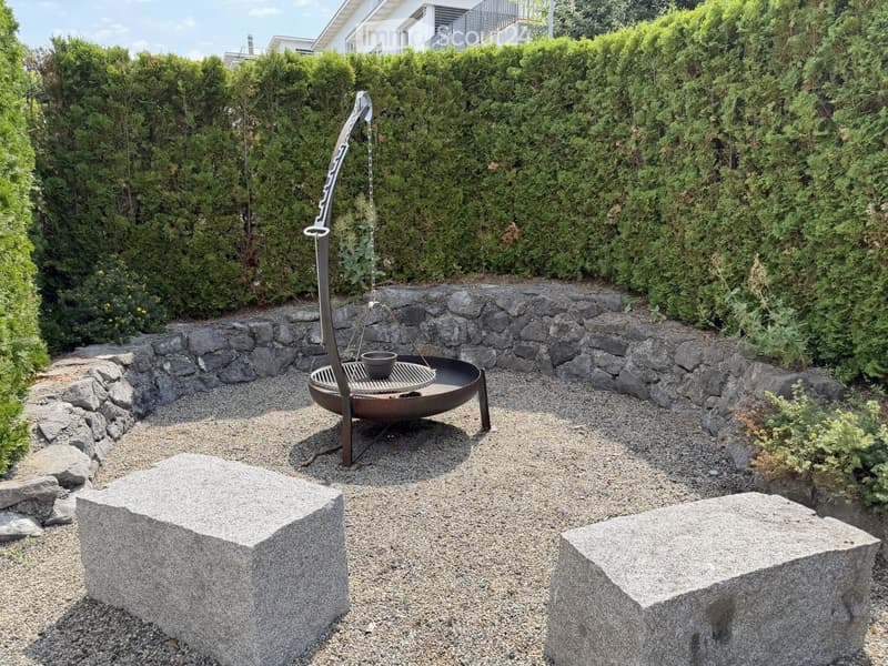 Garden area with a stone fire pit, two large stone blocks, gravel ground, and surrounding greenery.