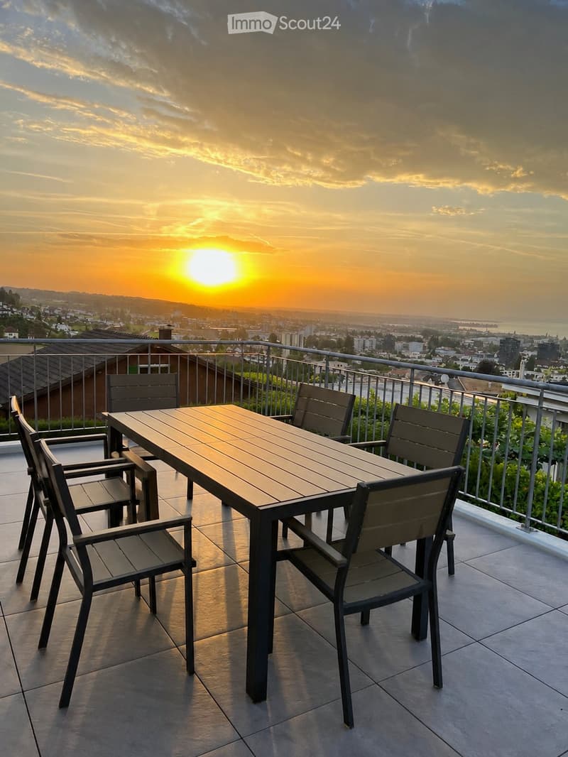 Balcony with a wooden table and four chairs, overlooking a cityscape at sunset.