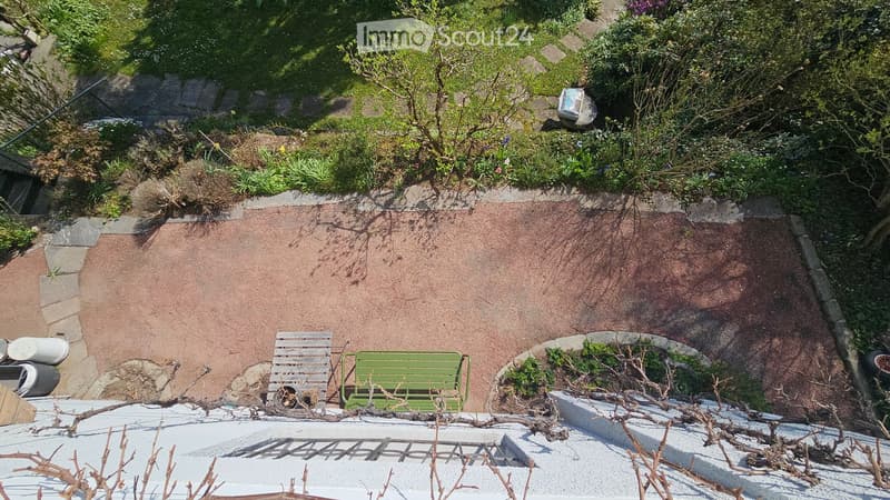 A small, elevated garden area with a reddish-brown ground cover, a green bench, a white chair, and surrounding greenery. The area is bordered by stone walls and has a small light fixture on the upper level.