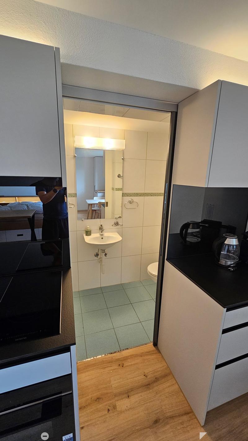A modern kitchen with a sink, mirror, and coffee maker, connected to a small bathroom with a toilet and sink.