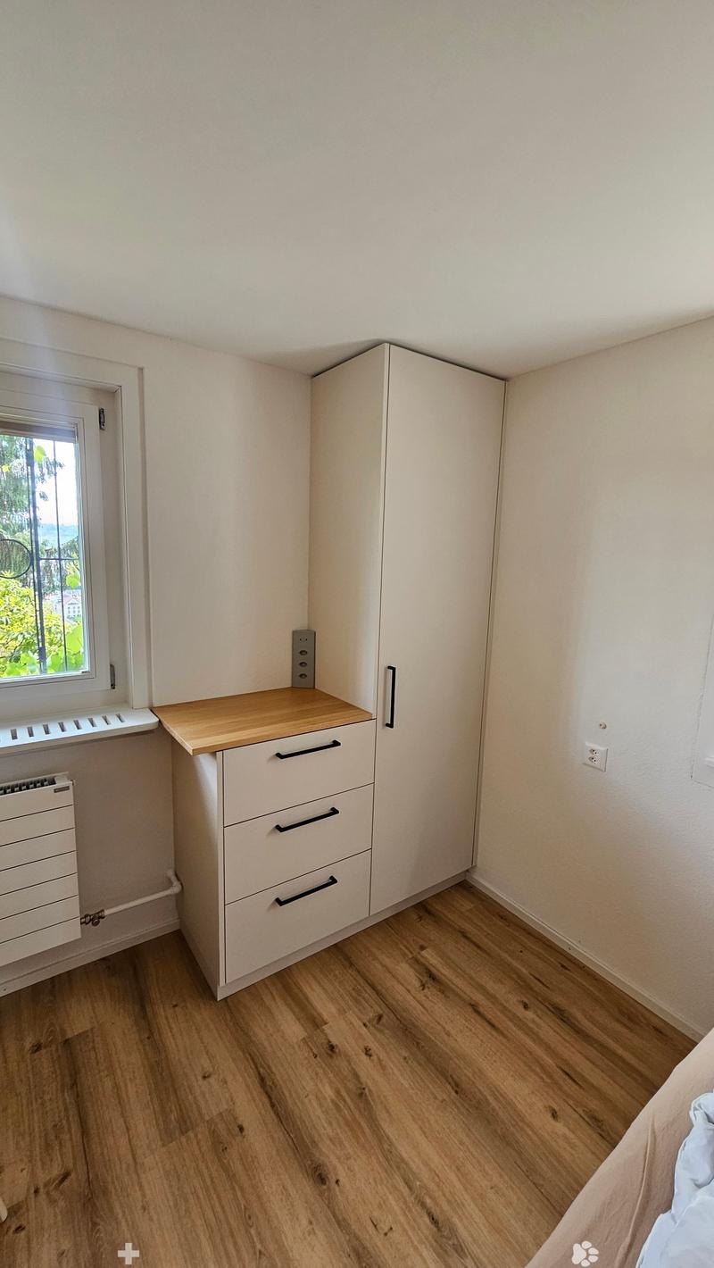 A small, well-lit bedroom with light wooden flooring, a bed, a wardrobe, a desk, and a drawer unit.