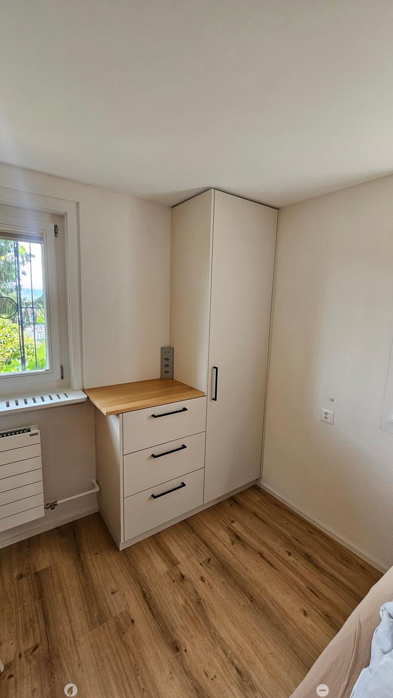 A small, well-lit bedroom with light wooden flooring, a bed, a wardrobe, a desk, and a drawer unit.