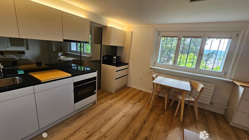 Modern kitchen with white cabinets, black countertops, and wooden flooring. Features a sink, oven, cutting board, small dining table with chairs, and a view through large windows.