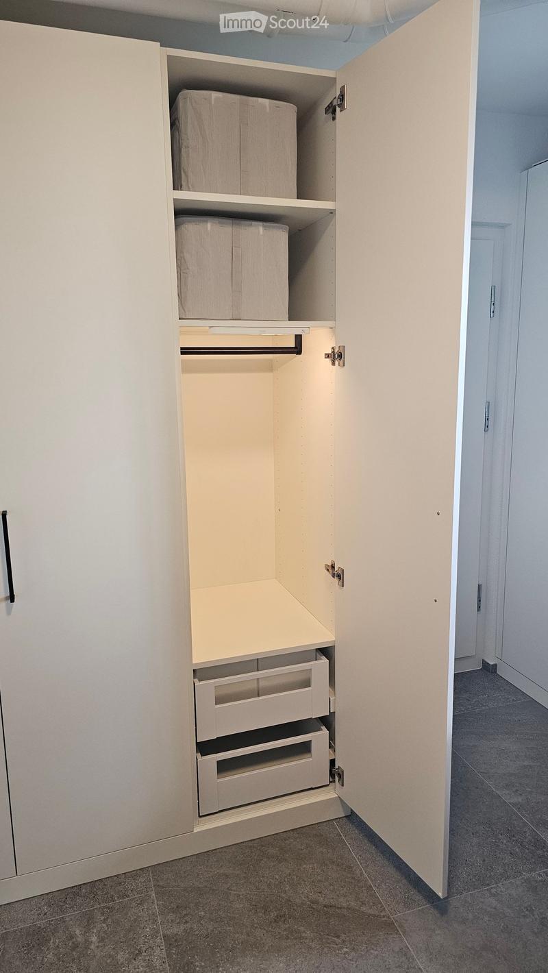 Open closet with white cabinetry, multiple shelves, and drawers, containing folded items. The hallway has tiled flooring and white walls.