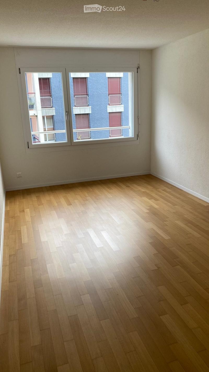 Empty room with wooden flooring and large windows showing a view of neighboring buildings.
