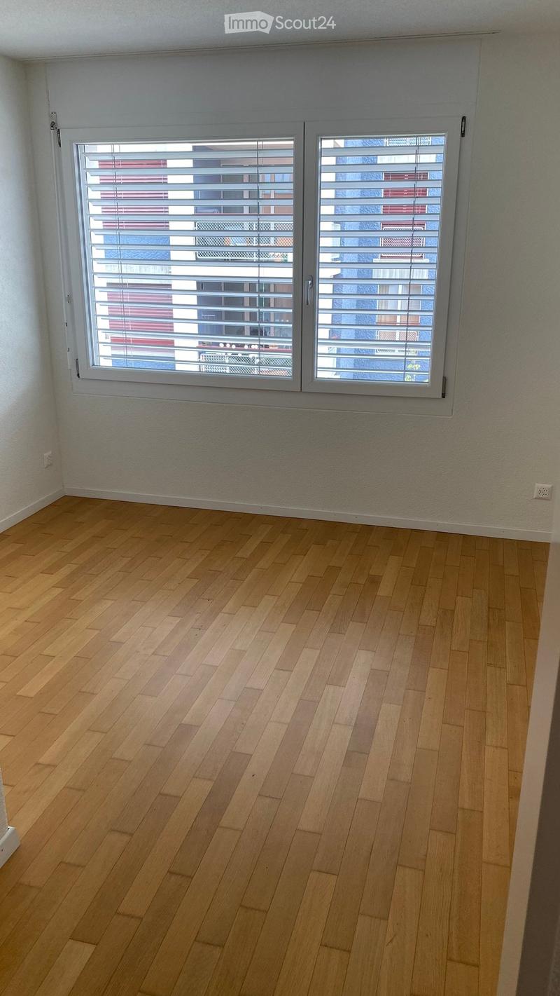 Empty room with wooden flooring and large windows with blinds, showing a view of neighboring buildings.