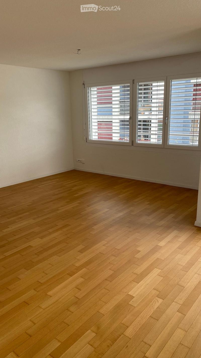 Empty room with light wooden flooring and large windows with blinds, showing a view of a building outside.
