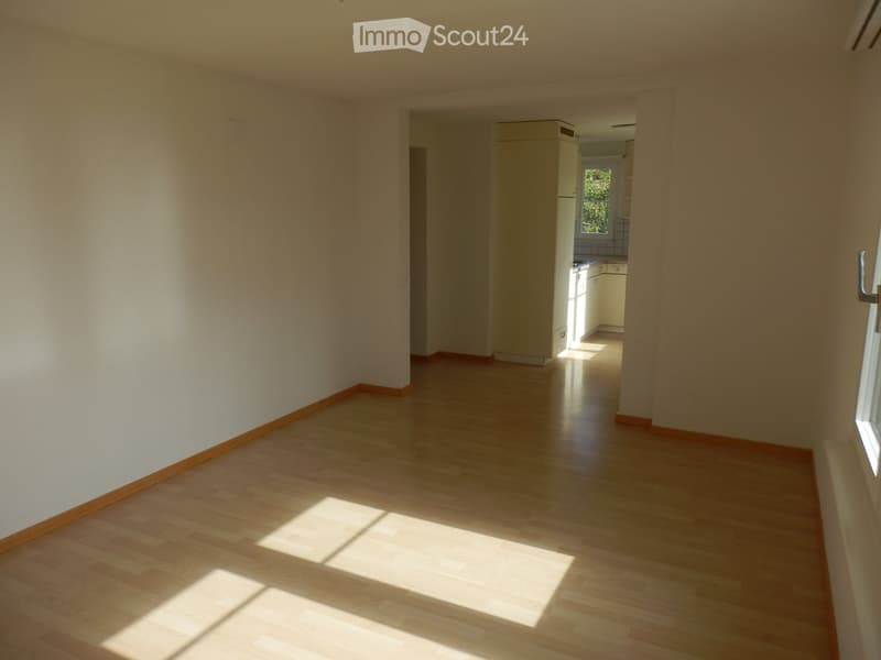 Empty hallway with light wooden flooring and white walls, leading to a kitchen area with cabinets and a window.