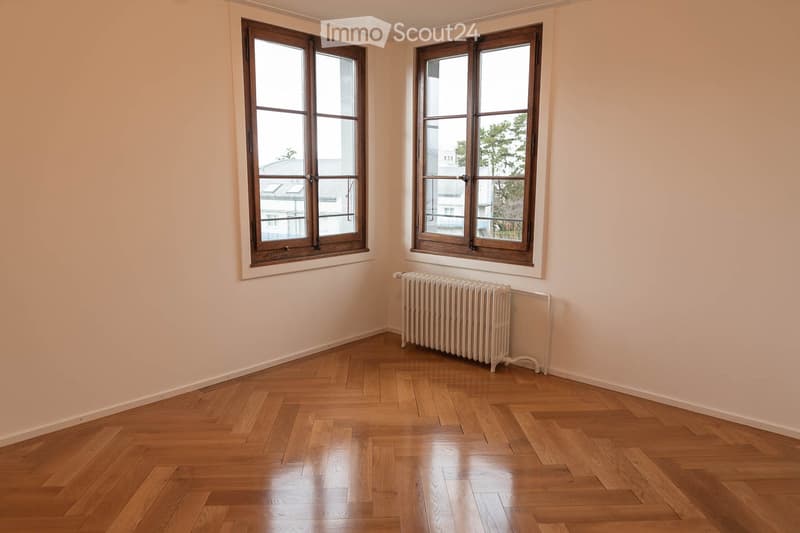 Empty room with wooden flooring, light-colored walls, and two large windows with wooden frames. A radiator is visible under one window.