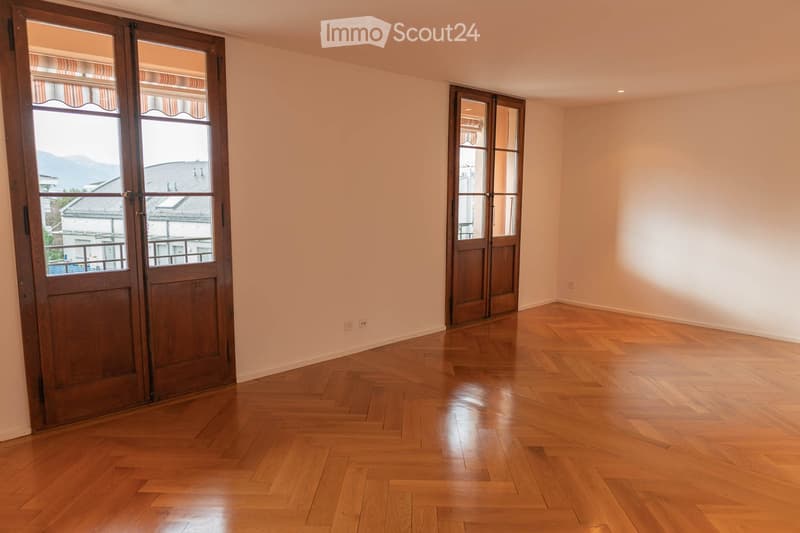 Empty room with wooden herringbone flooring, light-colored walls, and two large wooden doors with glass panels leading to a balcony.