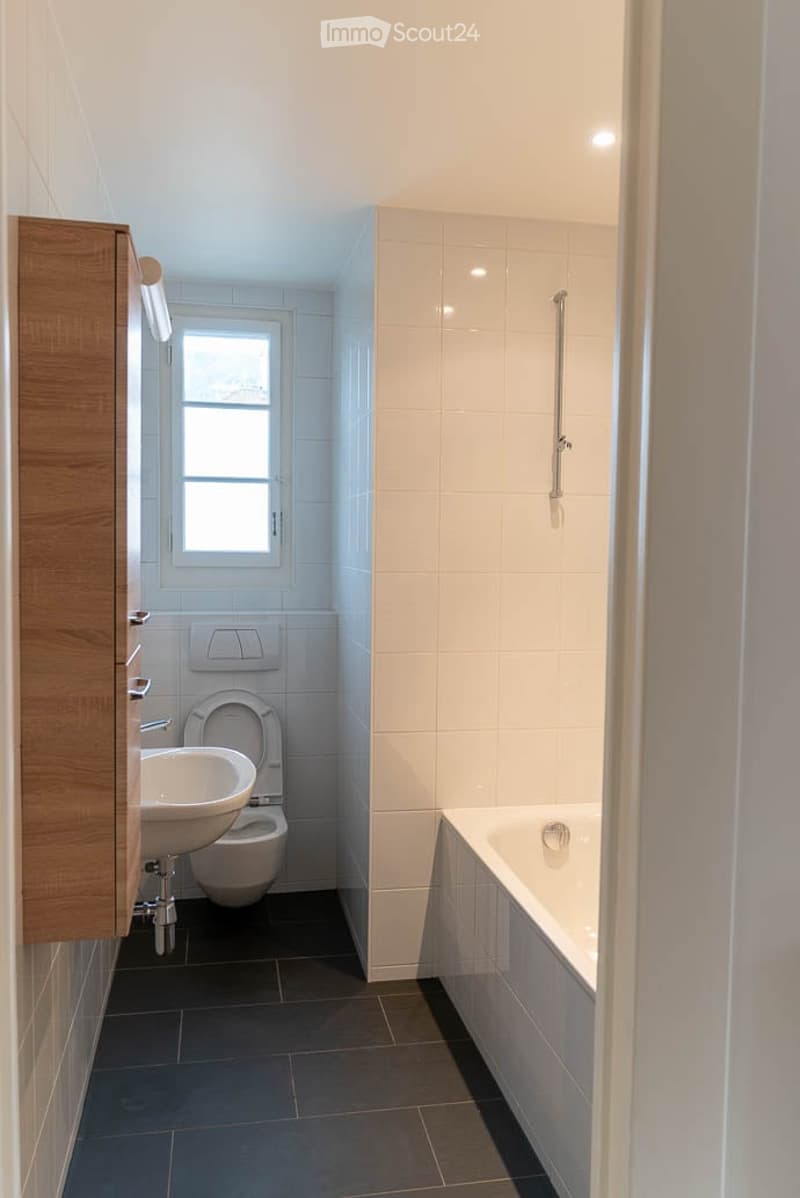 Modern bathroom with white tiles, dark grey floor, sink, toilet, bathtub, and a shower rail. A small window provides natural light.