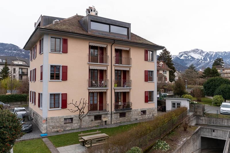 Multi-story residential building with beige facade, red shutters, balconies, and a scenic mountain view in the background.
