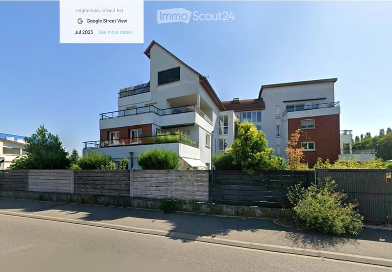 Modern multi-story residential building with balconies, surrounded by greenery and a wooden fence.