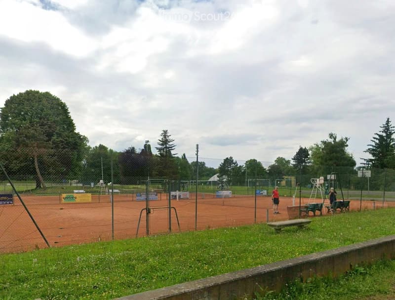 Outdoor tennis court area with fenced perimeter, grass, trees, and a few people visible in the distance.