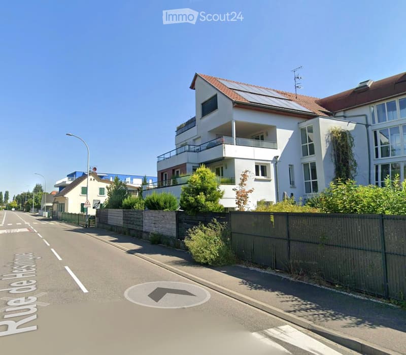 A modern two-story house with white walls, large windows, and a balcony on the upper floor. The house features a red-tiled roof and is surrounded by greenery and a wooden fence.